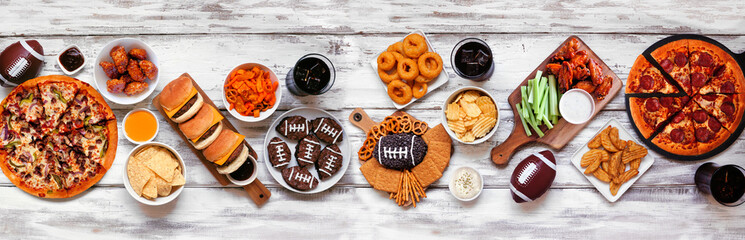 Super Bowl or football theme food table scene. Pizza, hamburgers, wings, snacks and sides. Top down view on a white wood background.