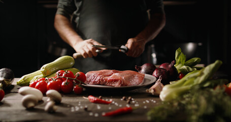 Chef sharpening his knife in front of kitchen table. Cooker preparing his tools before cutting raw piece of meat and various vegetables