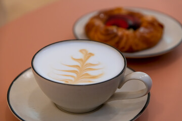 Close-up view of caffè latte with brown plant drawing in white porcelain cup standing on table in cafe. Blurred puff pastry cake with custard in the background. Selective focus. Coffee break theme. 