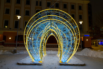 Yellow and blue Christmas lights arch decoration stands in night city. Copy space for your text or decoration. Selective focus. Template for people or product placement. Winter holidays theme.