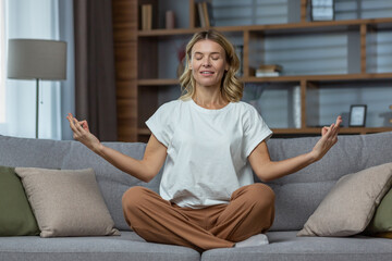 Senior woman at home resting meditating with eyes closed sitting on sofa in living room, housewife mature blonde in lotus pose smiling.