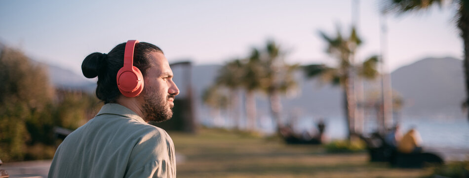 Portrait Of A Young Man In Bright Large Headphones Listening To Music, Audiobook By The Sea At Sunset.