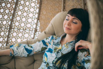 Portrait of a beautiful girl sitting in a round hanging chair. Cute brunette woman peacefully resting in armchair