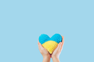 Handmade crochet amigurumi heart in the hands of a child on yellow and blue colors. Flag of Ukraine. Isolated on a blue background. Space for text.