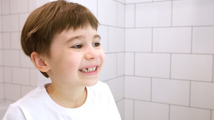 A child looks in the mirror at his beautiful smile. Handsome caucasian 6 year old boy in a white T-shirt on a white bathroom background. Happy mood. Lower incisor. Copy space