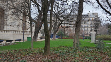 Un parc dans une zone urbaine, avec des bâtiments au fond, un gazon bien entretenu, beaucoup de vert, des arbres sans feuilles, du jardinage, avec des bancs peu occupés et peu de passants, ciel gris e - obrazy, fototapety, plakaty