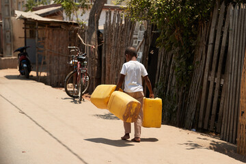 Malagasy boy carrying three yellow water cans, shot from behind. Poverty in Madagascar. Travel photo.