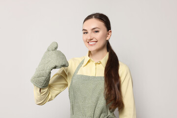 Beautiful young woman in clean apron with pattern and oven glove on light grey background