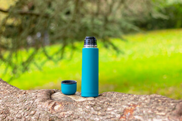 Steel green thermos and mug with hot tea on a branch of an old coniferous tree in the forest. Nature, picnic, camping