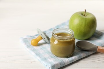 Jar with healthy baby food, apple, pacifier and spoon on white wooden table. Space for text