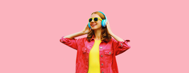 Portrait of happy smiling young woman in headphones listening to music wearing jacket on pink background