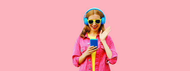 Portrait of stylish modern happy smiling young woman in headphones listening to music with smartphone wearing jacket on vivid pink background