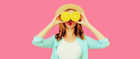 Summer portrait of cheerful young woman covering her eyes with slices of orange fruits and looking for something wearing travel straw hat on pink background