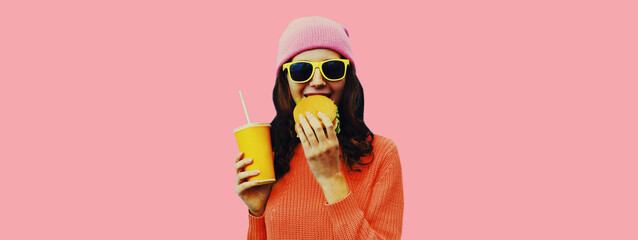 Portrait of stylish young woman drinking juice with burger fast food on pink colorful background