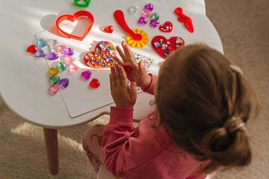Child hands creating red heart from play dough for modeling with decorate from crystal rhinestones and shiny stones. Toddlers crafts for Valentine's Day. Holiday Art Activity for Kids.