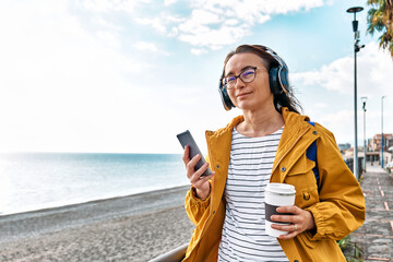 Woman with closed eyes listening music or podcast from smartphone application in headphones in nature. Smiling middle-aged female in headphones while walking near the sea.