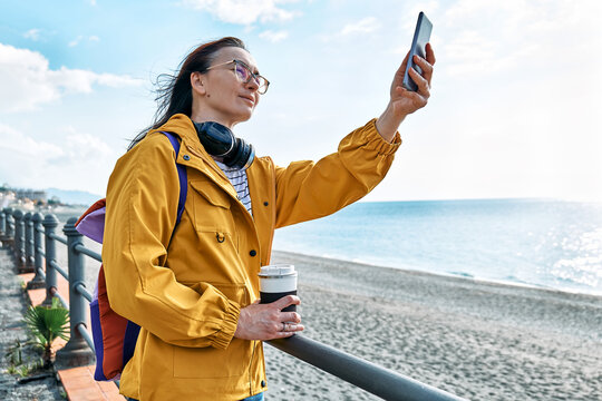 Woman with backpack taking photo of the sea on smartphone on seaside promenade. Relaxing outdoors. The concept of hiking and travel in the low season.