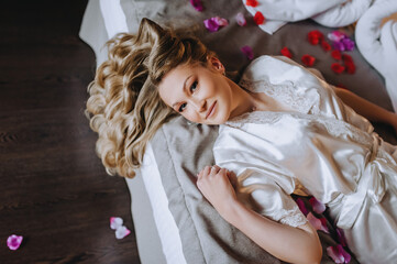 A beautiful smiling curly blonde bride in lingerie, a dressing gown lies in the morning in a room on a bed in rose petals, a bedroom. wedding photography.