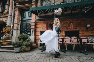 Obraz premium A beautiful curly smiling blonde bride, biker, lover of musical rock in a black leather jacket, white dress with a bouquet of reeds stands, dances in the city. Wedding photography, portrait.