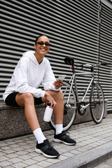 Fototapeta na wymiar Cheerful african american woman in sunglasses holding smartphone and bottle near bike on street 