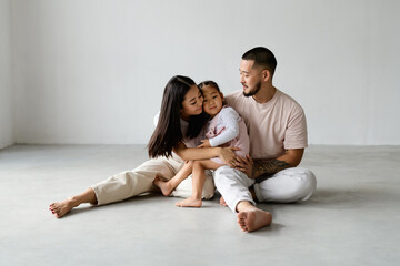 Young asian parents hugging toddler child together while sitting on grey background