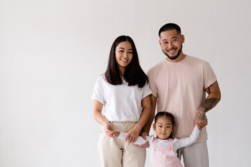 Fototapeta na wymiar Smiling young asian parents holding hands of toddler daughter and looking at camera isolated on grey 