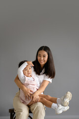 Positive asian mom looking at camera and hugging toddler daughter on grey background 