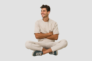 Young caucasian man sitting on the floor isolated on white background smiling confident with crossed arms.