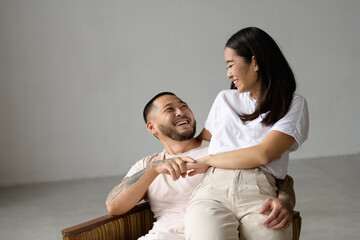Tattooed asian husband holding hand of cheerful wife on armchair on grey background 