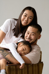 Portrait of cheerful asian parents looking at camera while hugging toddler daughter on armchair isolated on grey background 