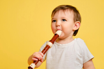 Toddler baby plays the flute, a child with a wind musical instrument on a studio yellow background....