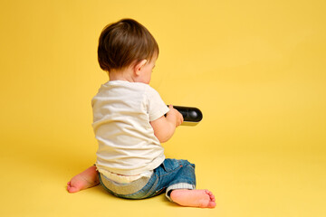Toddler baby plays with a wireless music speaker on a studio yellow background. Happy child in a...