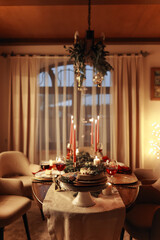 Festive table setting for celebration Christmas and New Year holidays in living room. Dining table with traditional dinner meal decorated burning candles, fir branches, xmas baubles, holiday garlands