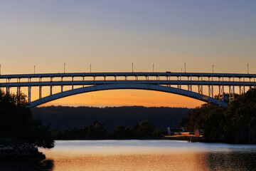 Fototapeta na wymiar Henry Hudson Bridge, a steel arch bridge, connecting the Bronx to Manhattan at sunset with a clear, glowing blue and orange sky and rippling water of the Hudson River passing under, with copy space