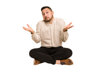 Adult latin man sitting on the floor cut out isolated doubting and shrugging shoulders in...