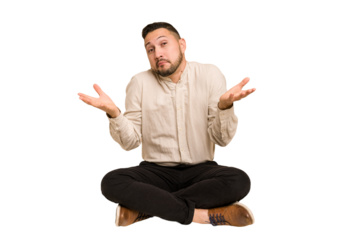 Adult latin man sitting on the floor cut out isolated doubting and shrugging shoulders in questioning gesture.