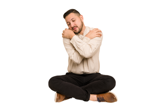 Adult latin man sitting on the floor cut out isolated hugs, smiling carefree and happy.