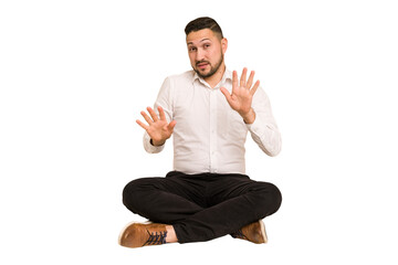 Adult latin man sitting on the floor cut out isolated rejecting someone showing a gesture of...
