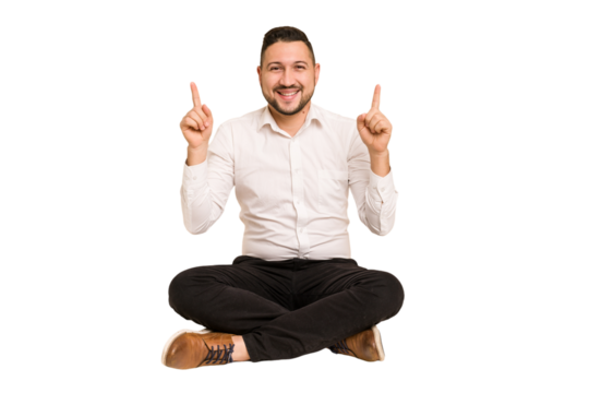Adult latin man sitting on the floor cut out isolated indicates with both fore fingers up showing a blank space.