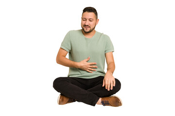 Adult latin man sitting on the floor cut out isolated touches tummy, smiles gently, eating and...