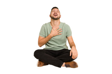 Adult latin man sitting on the floor cut out isolated laughs out loudly keeping hand on chest.