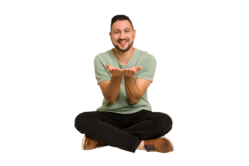 Adult latin man sitting on the floor cut out isolated holding something with palms, offering to camera.