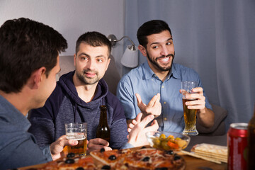 Three young glad men talking and laughing while enjoying beer and pizza at home