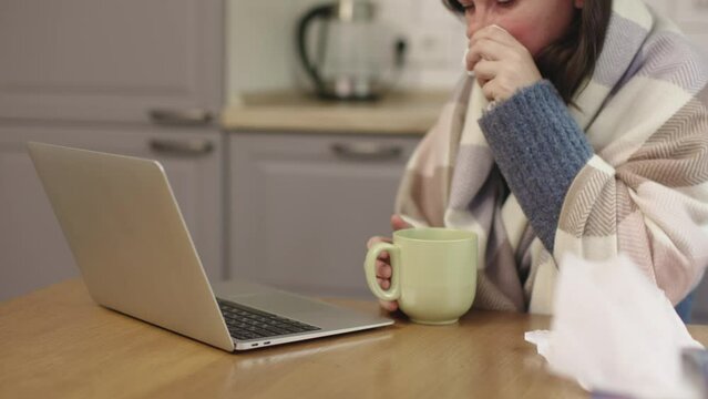 sick woman wrapped warm blanket using tissues for running nose working laptop sit at kitchen table cold indoors turn off heat economy problems gas supply 