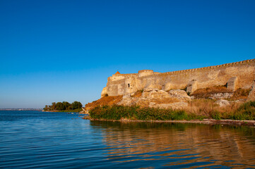 Fototapeta na wymiar Old stone fortress on the shore of the lake. Beautiful sunny landscape with fortress and blue sky