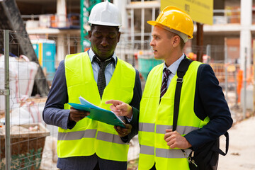 Two architects, Caucasian and African-american men in warnvests, standing on building site and discussing project documentation.