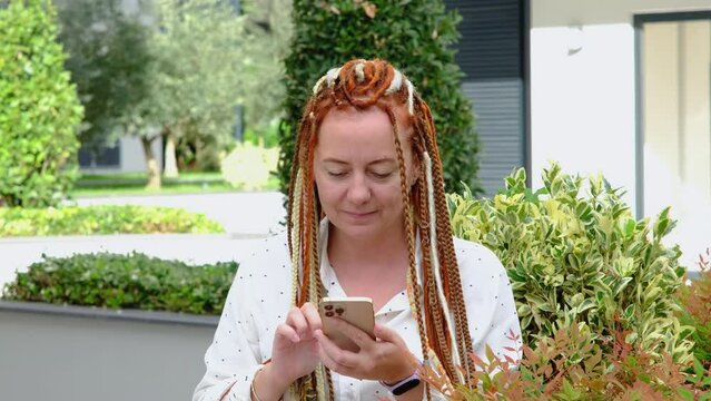 Modern confident young woman with red dreadlocks using mobile phone outdoor.