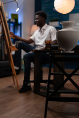 Artistic young man making artwork in home drawing studio with creative tools. Close up of vase used to create pencil masterpiece. Young african american artist sketching on easel at atelier.