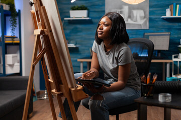 African american professional artist creating digital copy on electronic tablet of pencil masterpiece, sitting at creative workplace. Woman with experience sketching reviewing drawing on canvas.