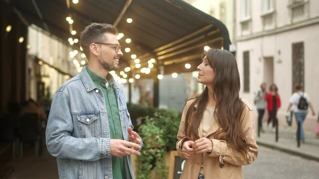 Side-view portrait of young beautiful cheerful man and woman talking standing in city center. Attractive successful male and pretty affable female having conversation wearing casual clothes.
